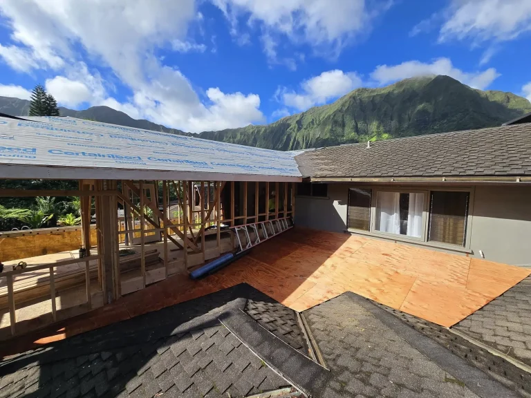 Wall framing and roof sheathing in progress beside an existing home with green mountains behind